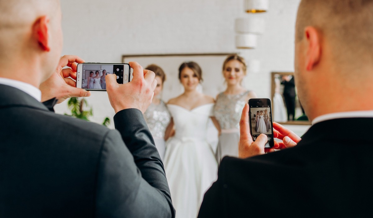 two men in suits take pictures of bride and two female wedding guests with smartphones