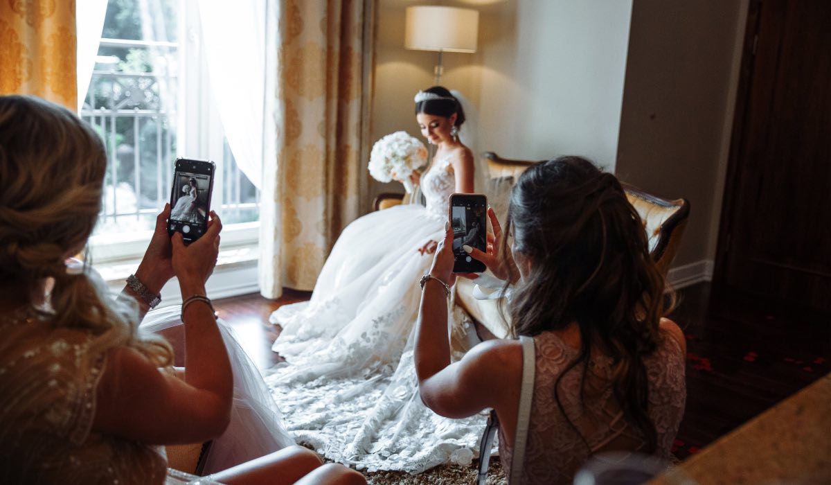 two women taking smartphone pictures of the bride sitting down next to a window