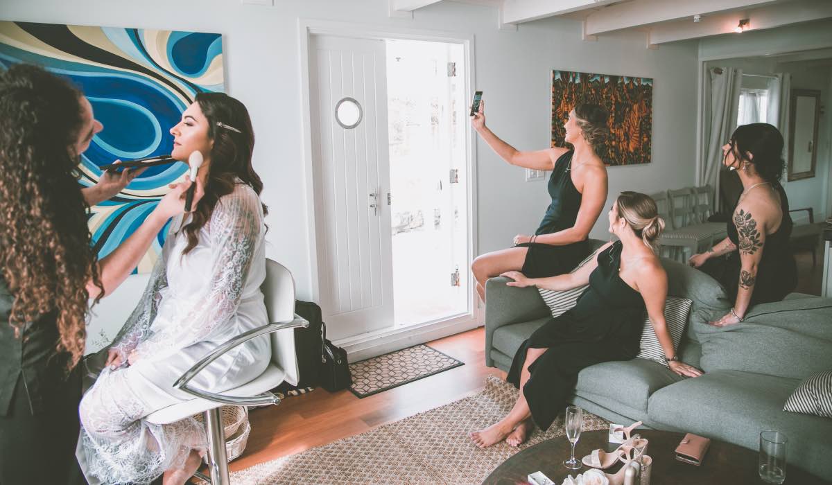 Bride is having her makeup done while bridesmaids are taking selfies