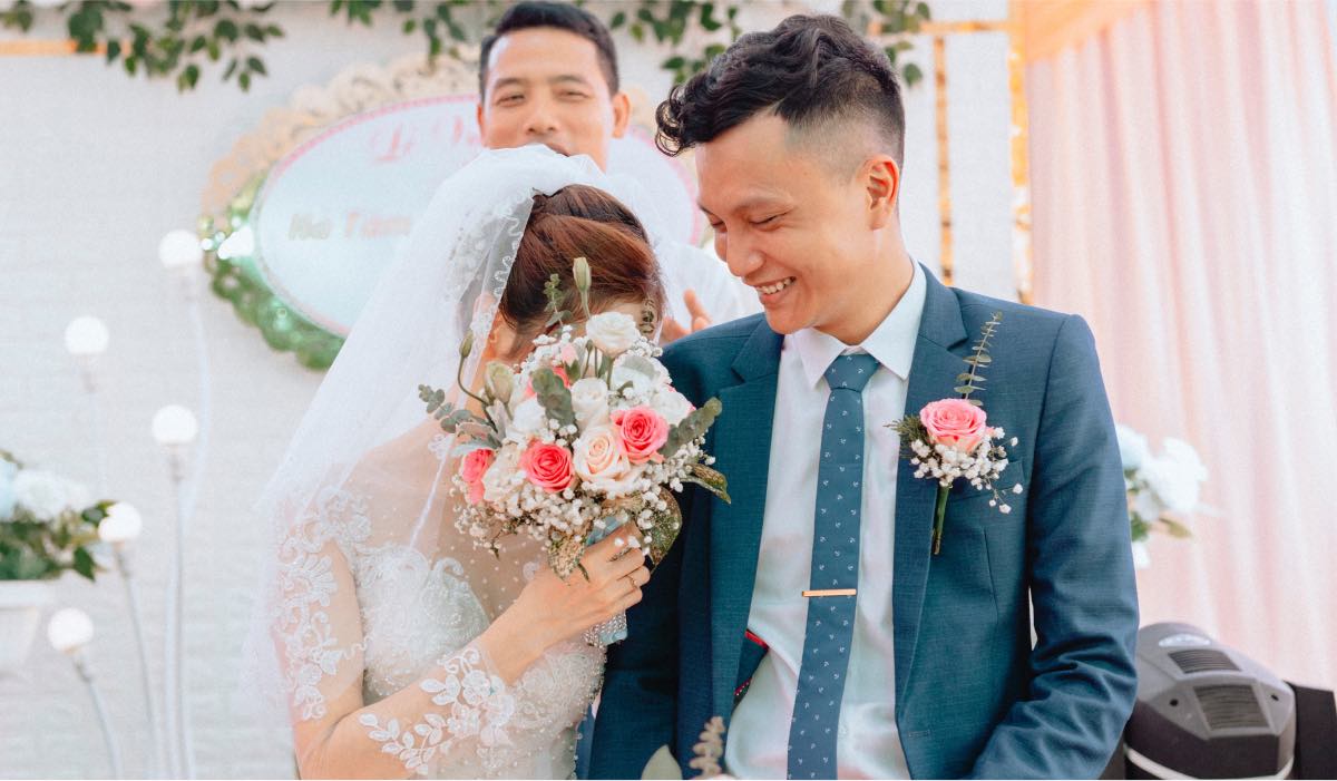 Groom embracing the bride while smiling down at her. The Bride is hiding her face behind a bouquet of flowers.